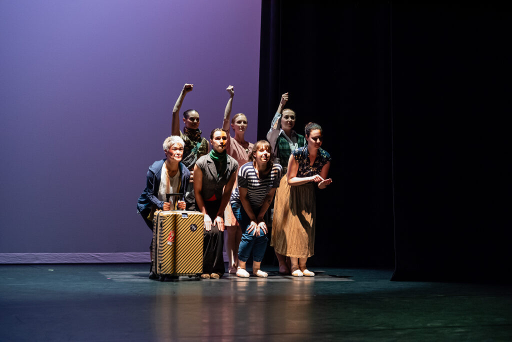 A group of dancers face forward, one holding a suitcase in front of her.