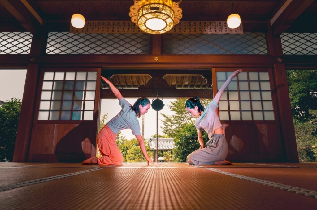 Two female dancers kneel, facing each other, one arm raised behind, in a temple with a garden in the background.
