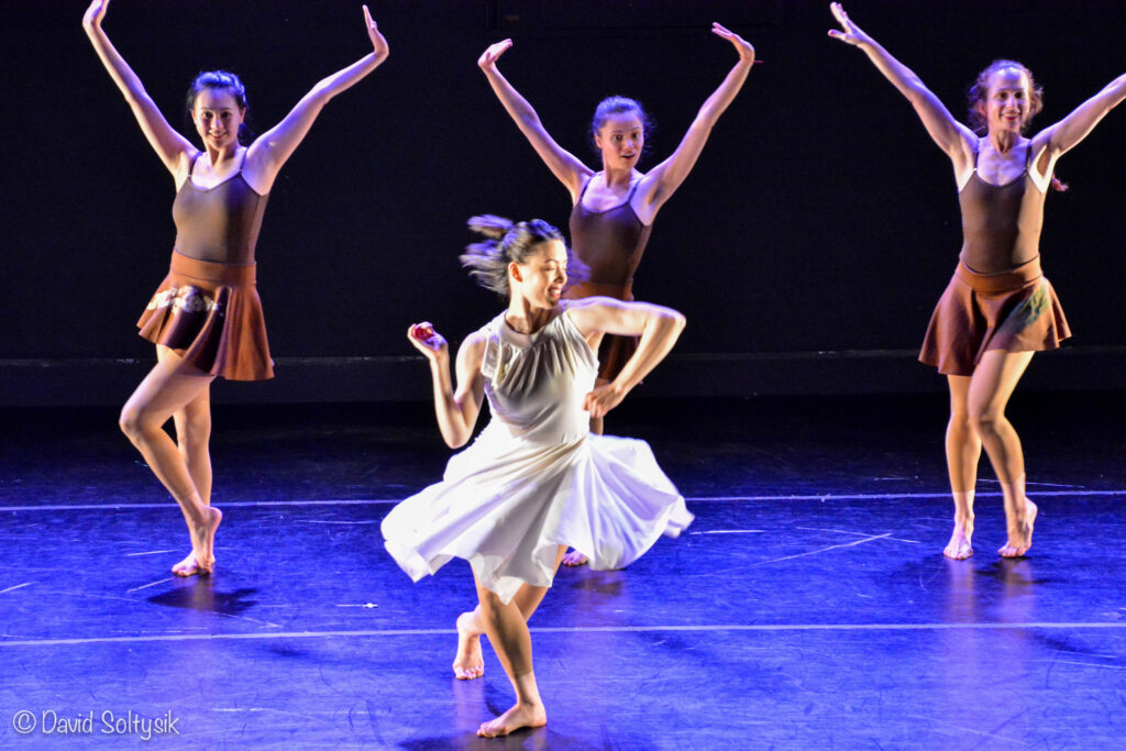 A female dancer in a white dress twists in front of three female dancers in brown.