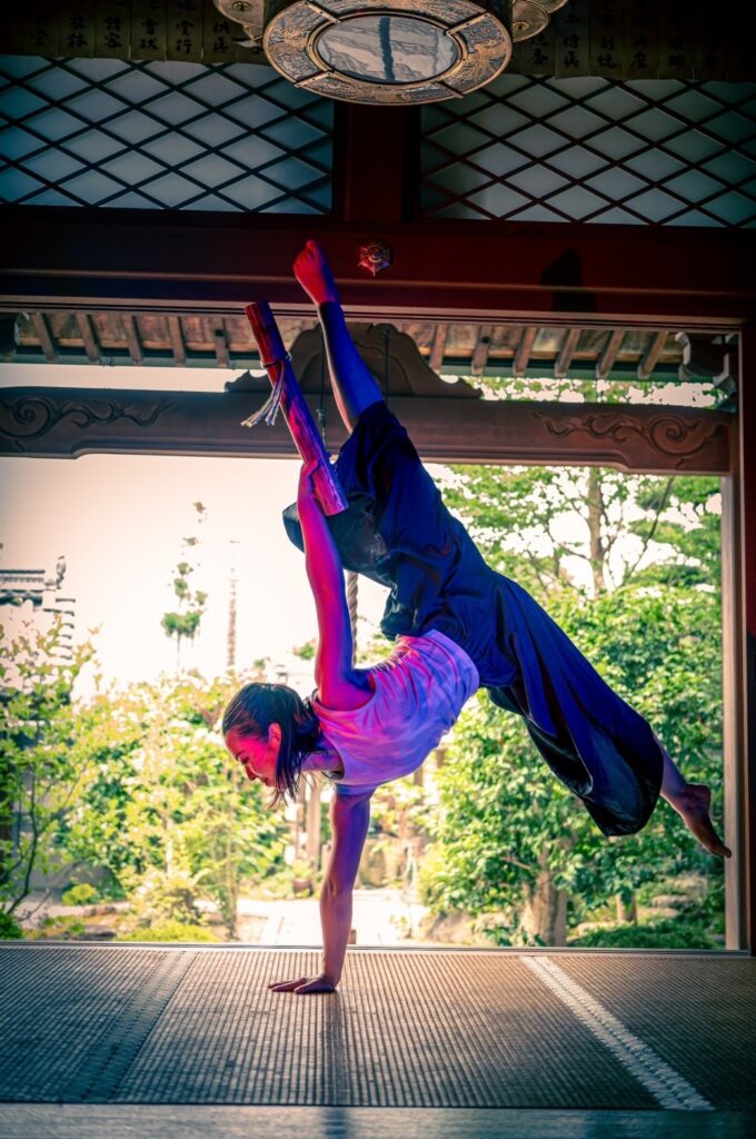 A female dancer balances on one hard, legs in a split, holding a rain stick in a temple with a garden in the background.