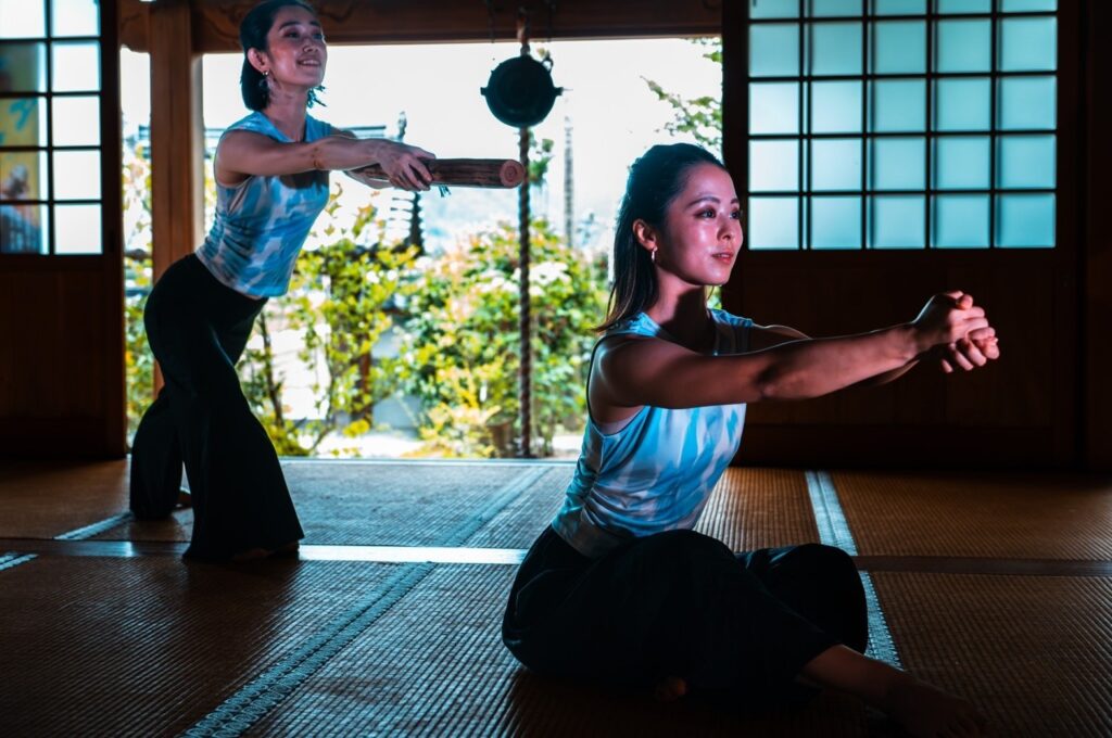 Two female dancers, one standing holding a rain stick, one seated with hand clasped in front of her, dance in a temple with a garden in the background.