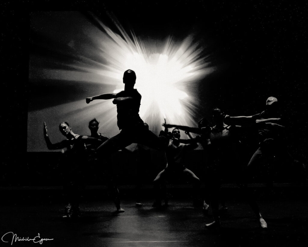 A backlit dancer, mid-jump, accompanied by other dancers.